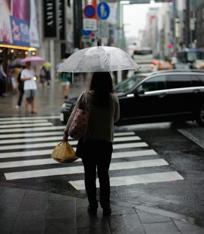 Fußgängerin mit durchsichtigem Regenschirm wartet an Zebrastreifen während schwarzes Auto passiert